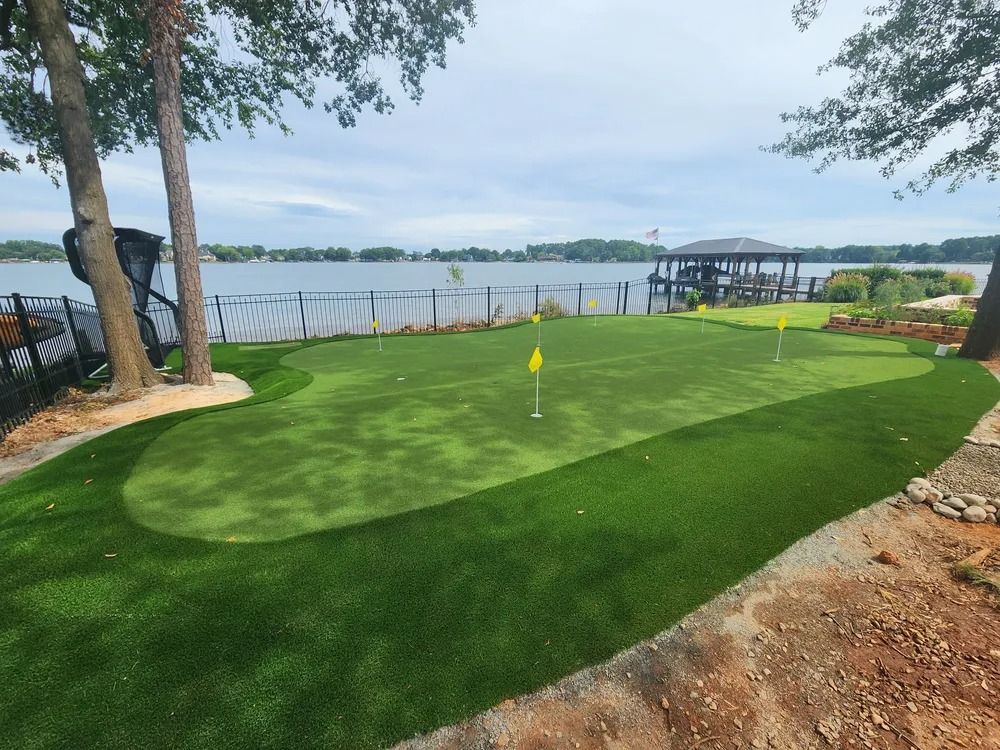 Waterfront putting green with flag, bordered by trees, dock, and a covered seating area.