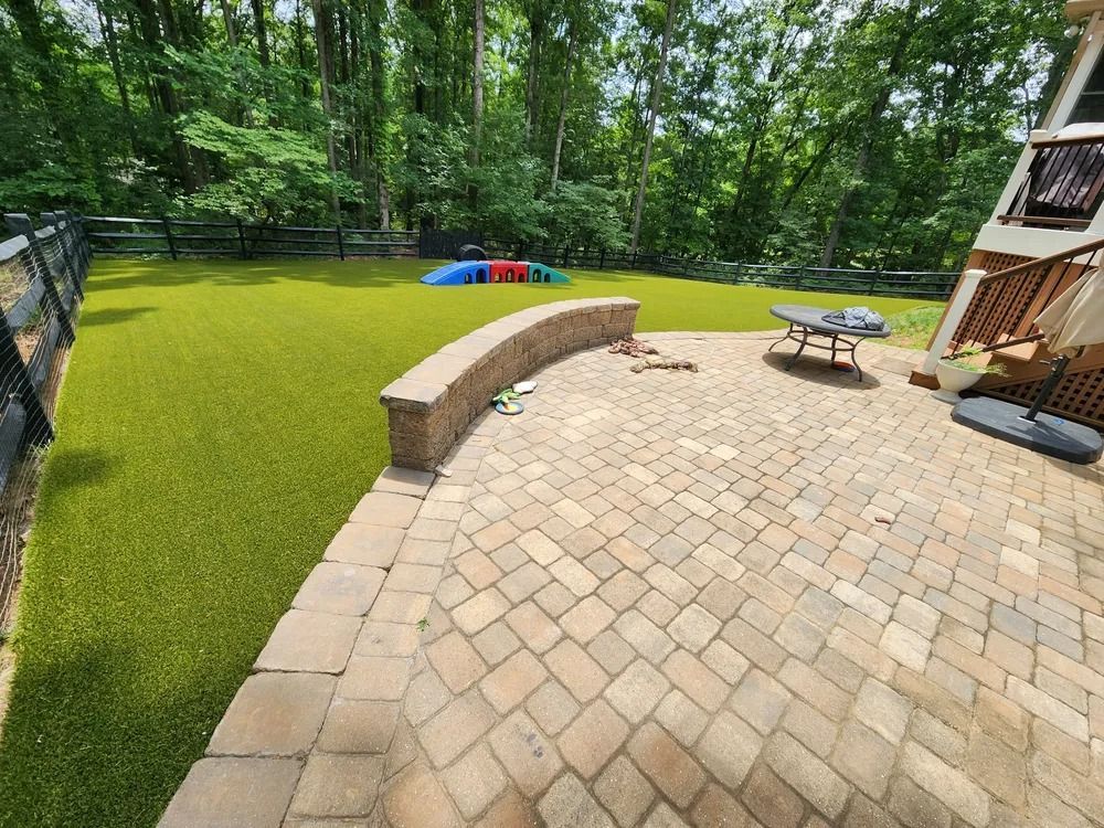 Backyard patio with curved stone bench, picnic table, artificial turf, and trees beyond fence