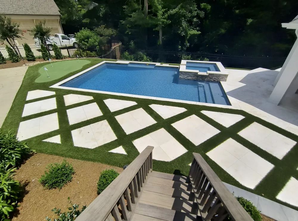 Backyard pool with geometric white pavers set in grass, stairs in foreground, and trees in the background