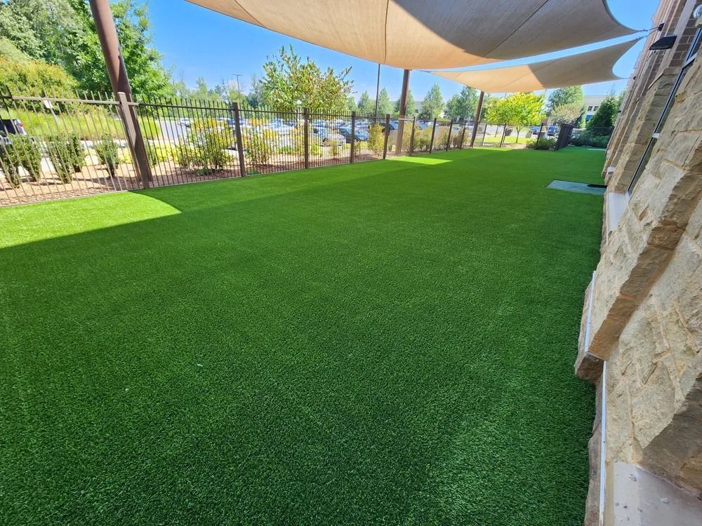 Shaded artificial grass courtyard with trees and stone walls beside a building