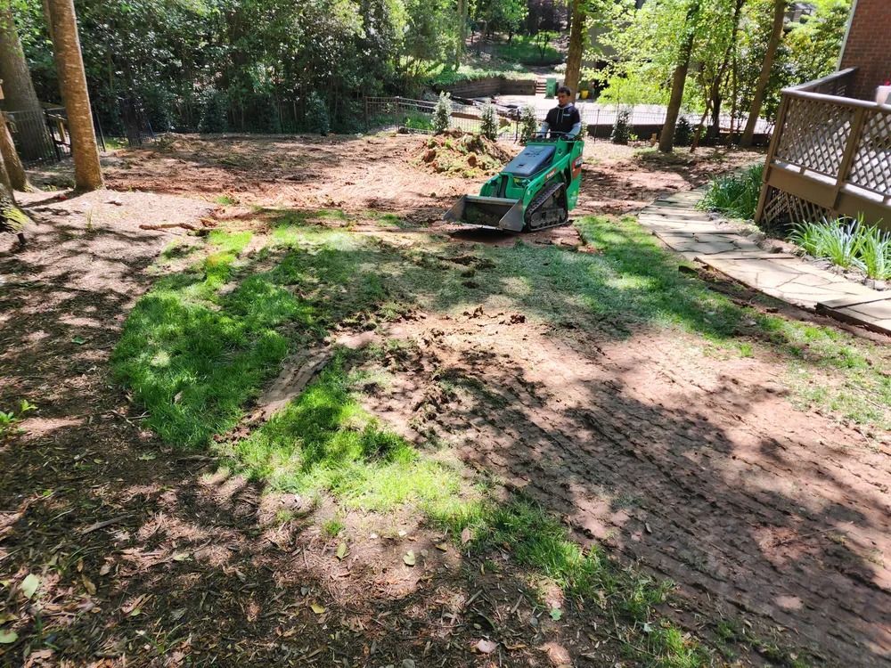 Dirt yard with a green lawn mower on a patchy lawn near trees and a wooden deck