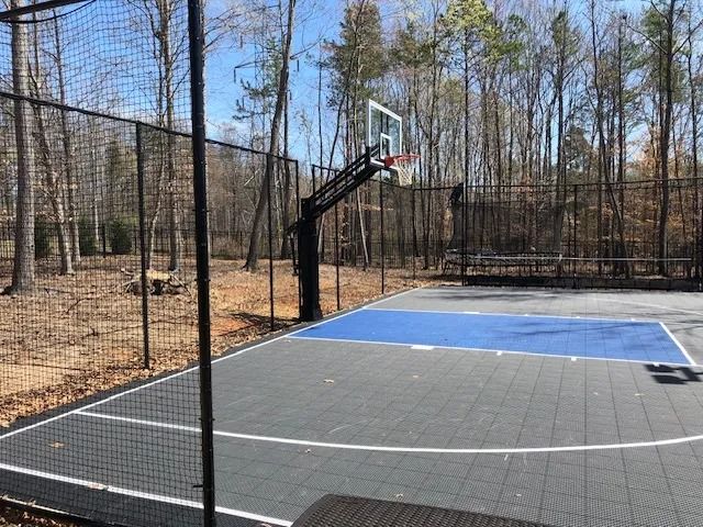 Outdoor basketball court with a blue-and-gray surface, hoop, and black fencing in a wooded area