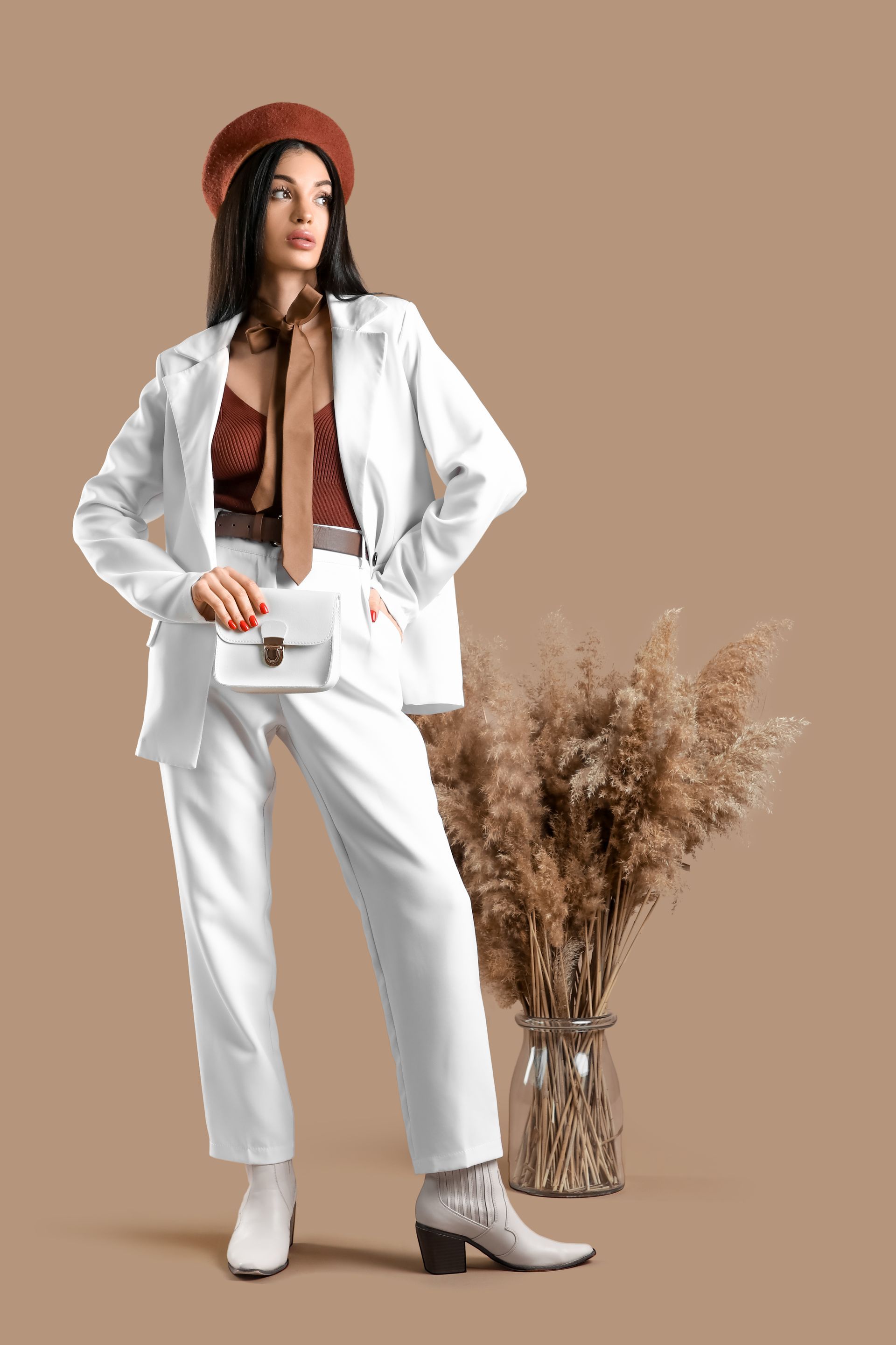 Woman in white suit and red beret posing beside dried pampas grass on a beige backdrop