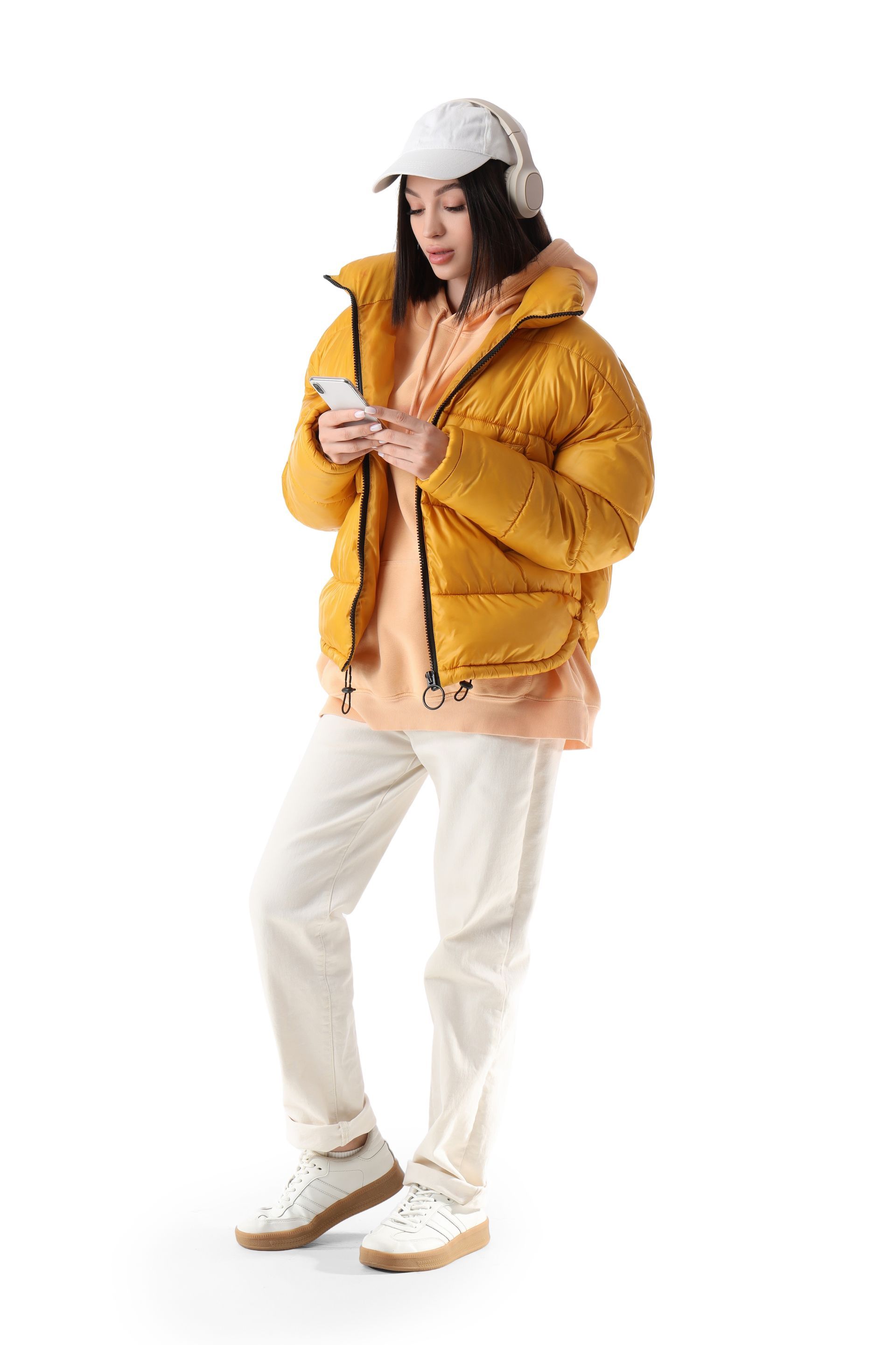 Person in mustard puffer jacket, cream pants, and cap looking at a phone against a white background