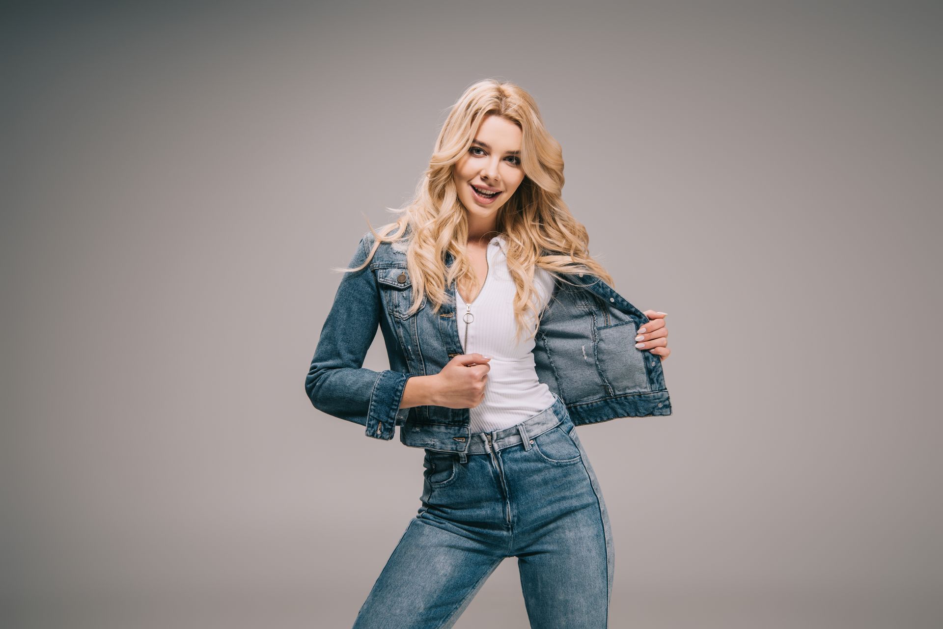 Blonde woman in denim jacket and jeans posing against a plain gray background