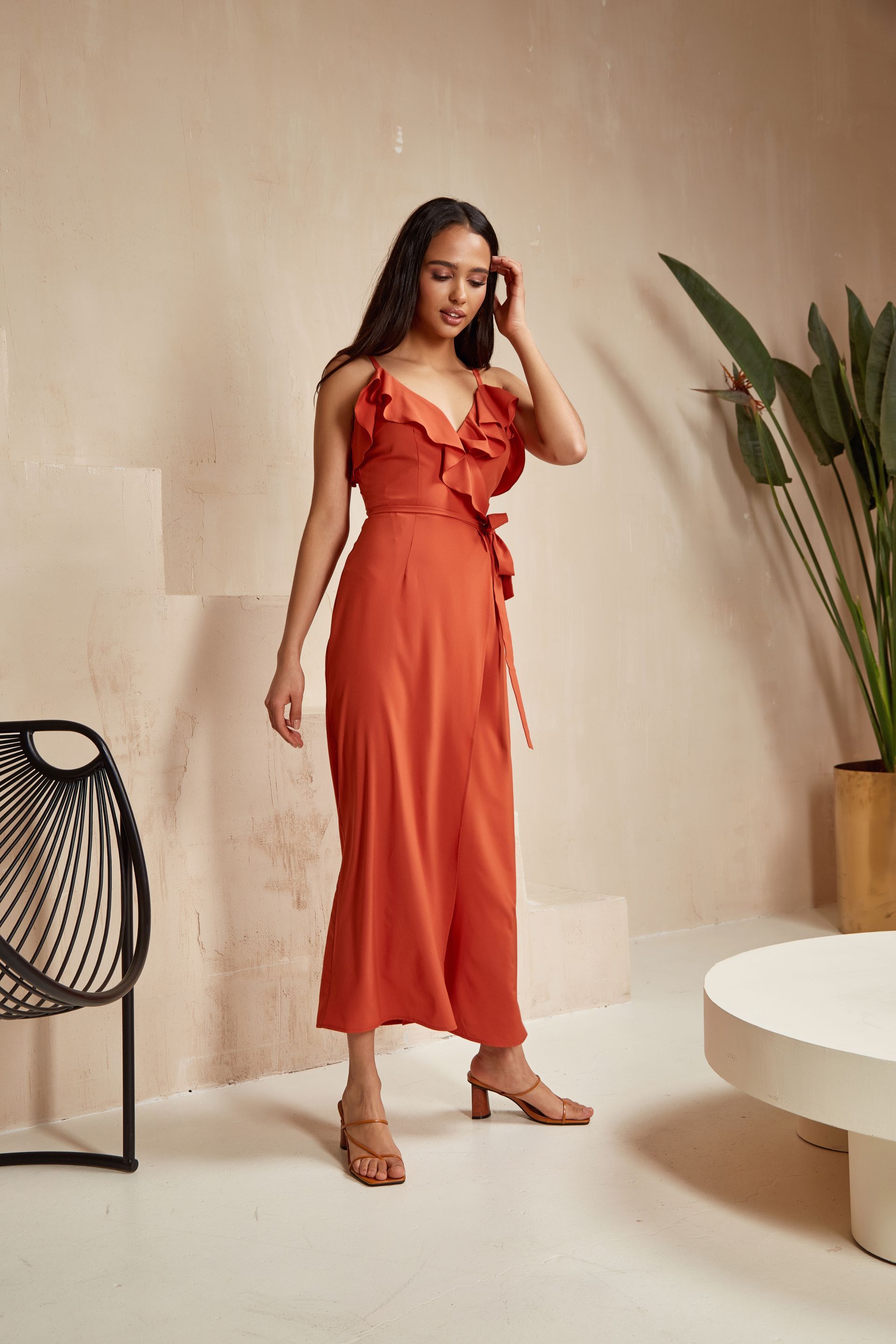 Woman in a rust-orange dress standing in a minimalist room with a beige wall, chair, plant, and round table.