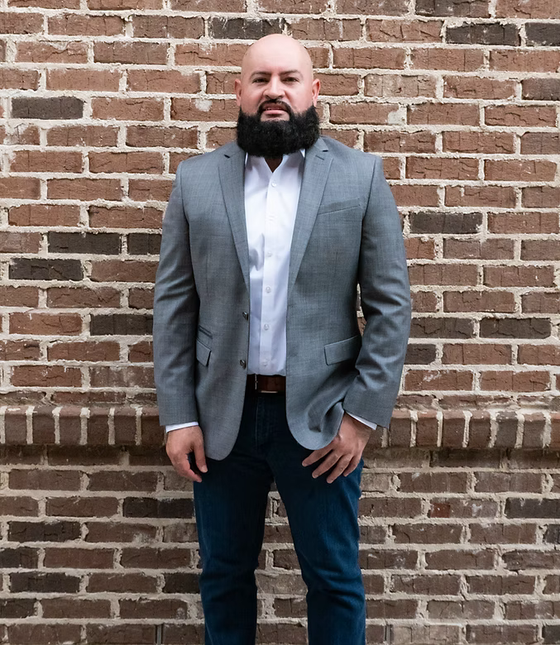 Man in gray blazer and white shirt standing against a brick wall.