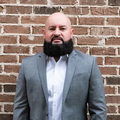 Man in a gray suit and white shirt standing against a brick wall, looking at the camera.