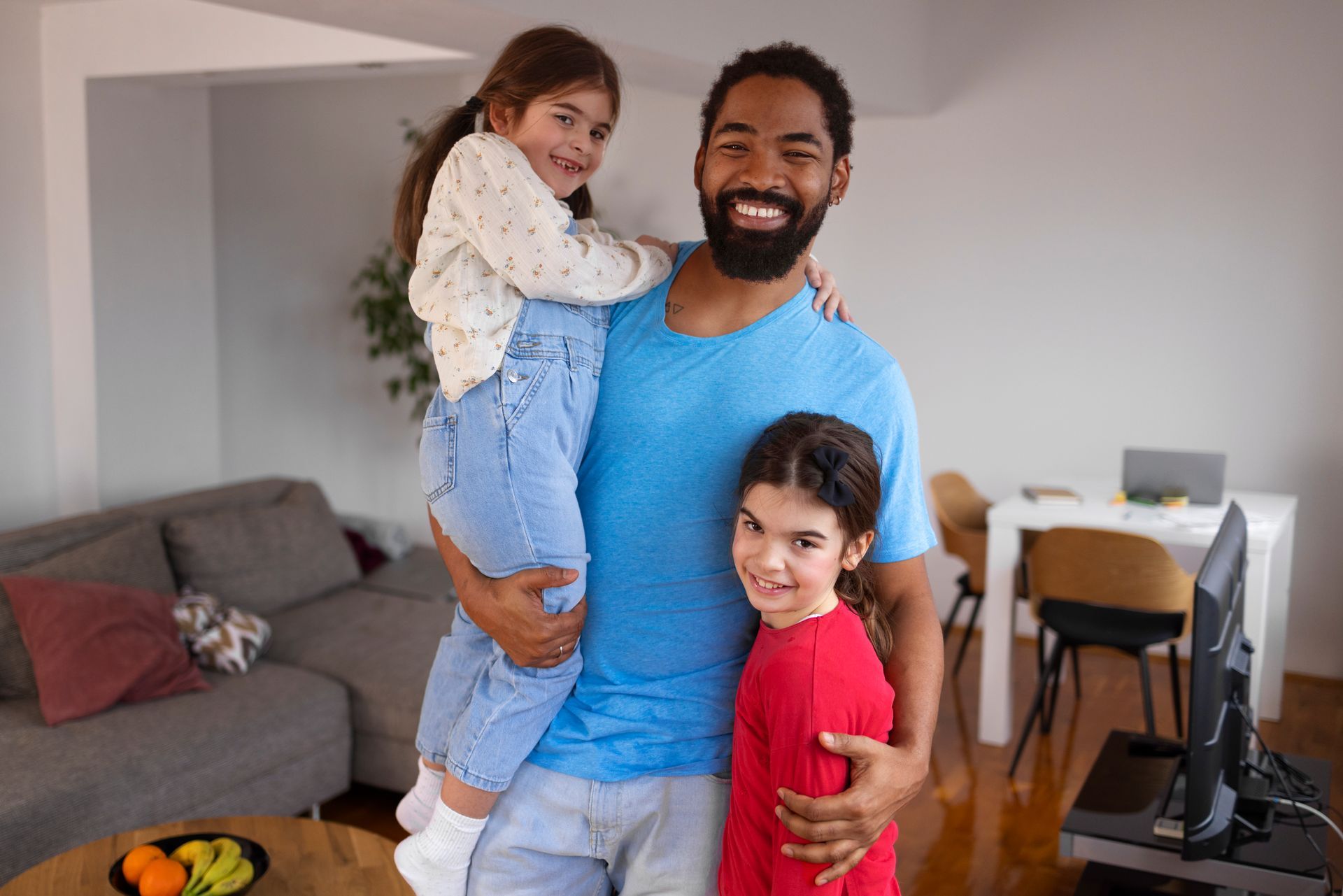 Three people smiling in a living room, one adult holding a child and another child standing beside them.