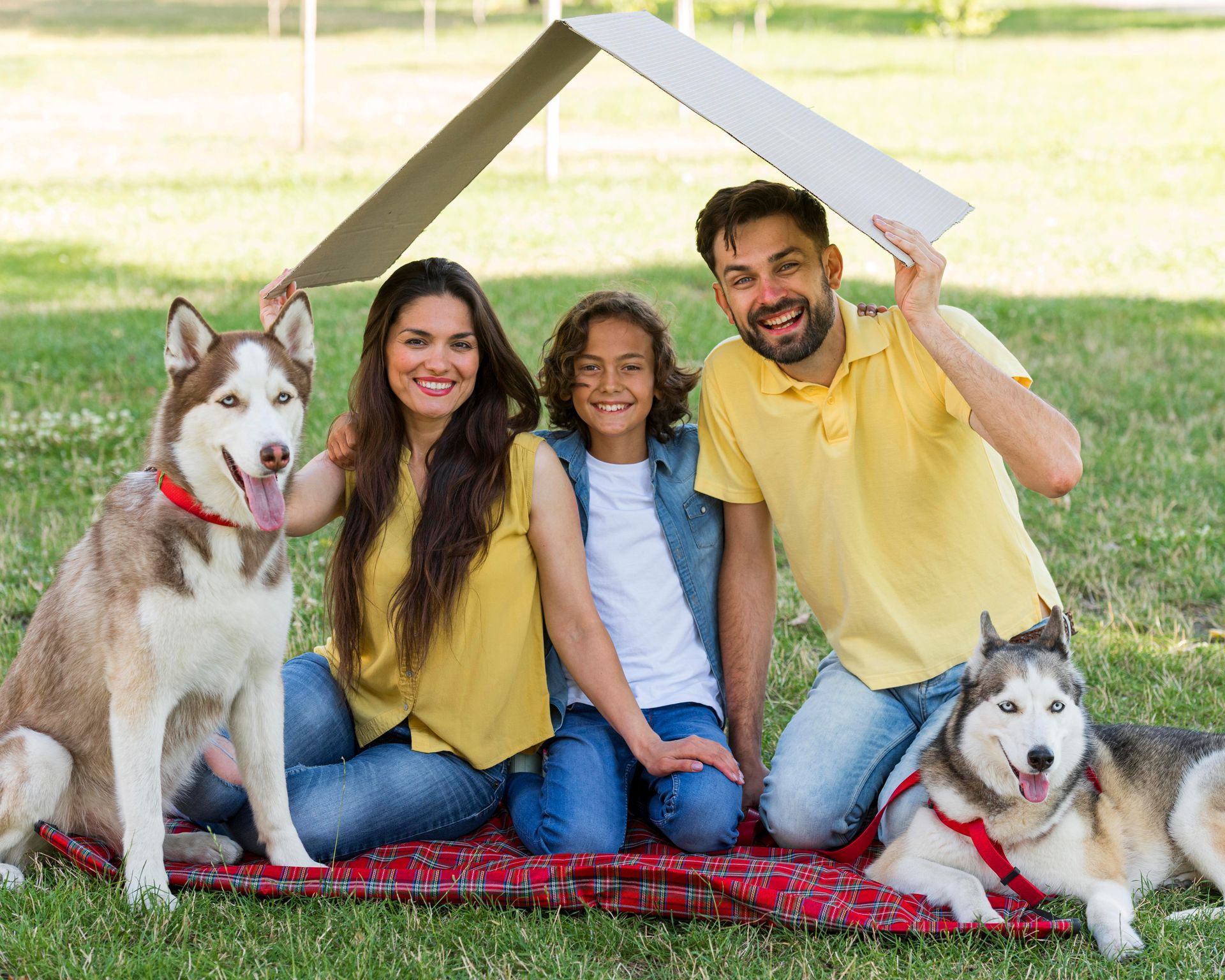 Family with two dogs posing on a lawn, smiling under a roof-shaped prop made of boards.