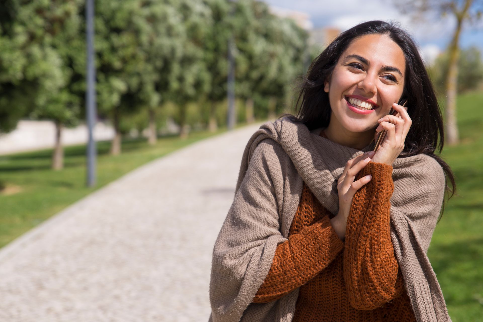 Smiling woman in a brown sweater standing on a park path beside green trees