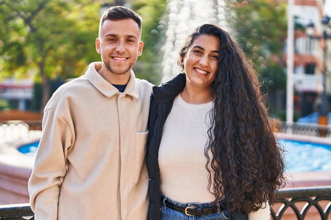 Two smiling people standing outdoors near a fountain, with trees and buildings in the background.