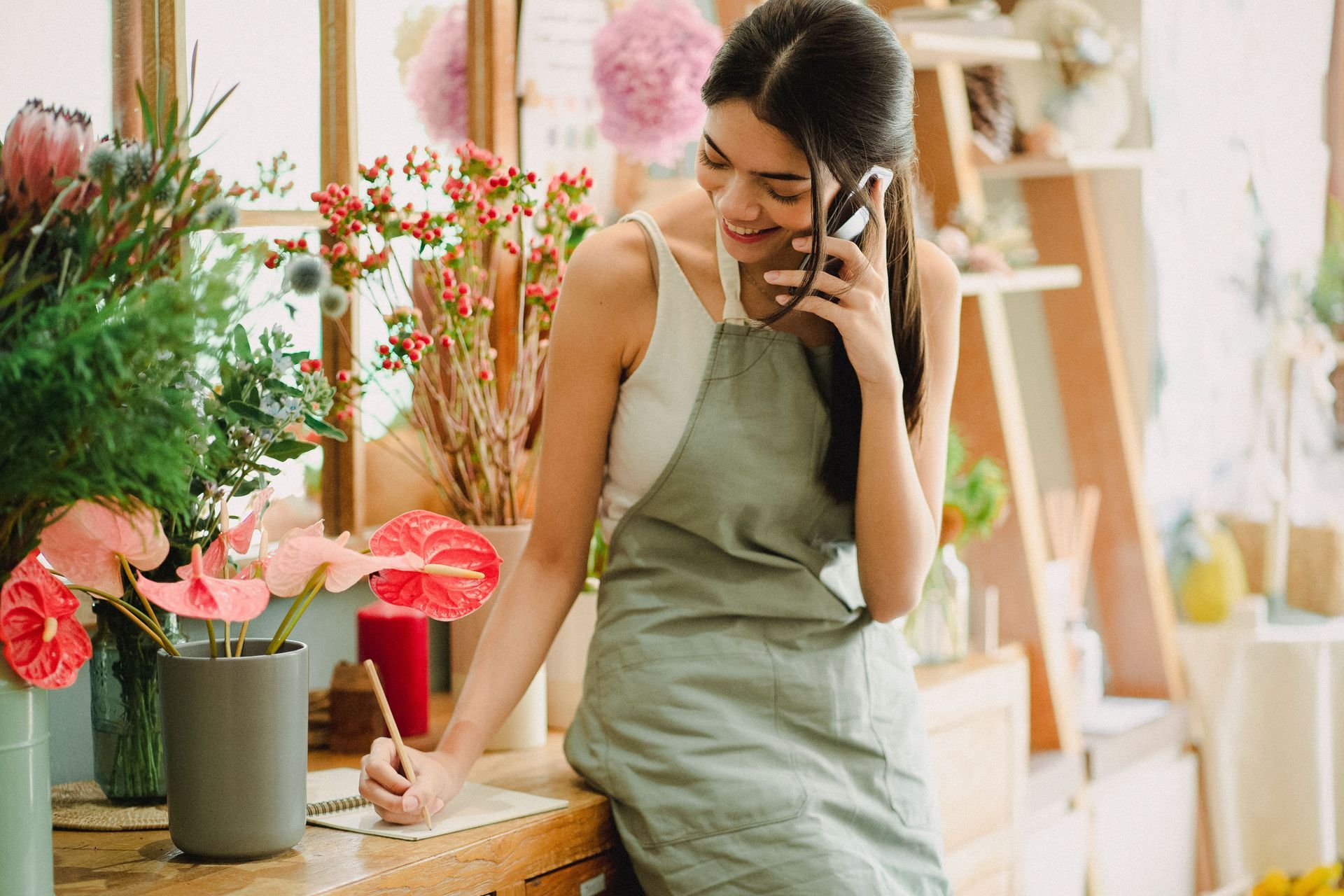 Woman in green apron smiling on the phone in a flower shop beside colorful bouquets