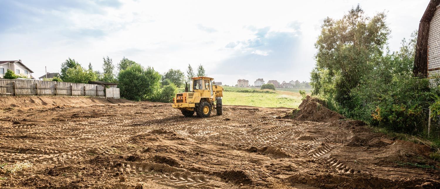 A yellow construction tractor levels a field of bare dirt on a bright, partly cloudy day.