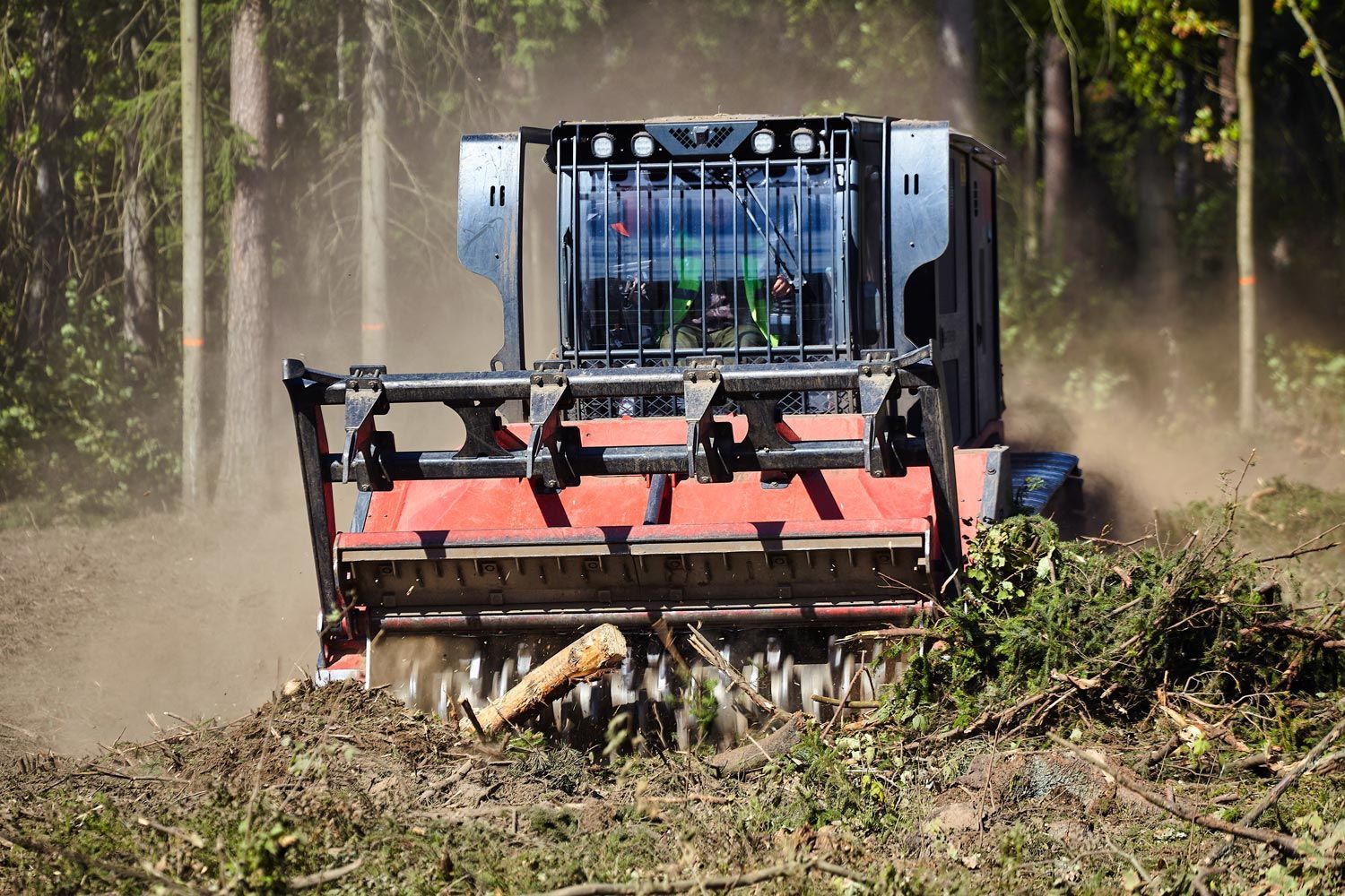 A red forestry mulcher clears brush and woody debris in a wooded area, kicking up dust.