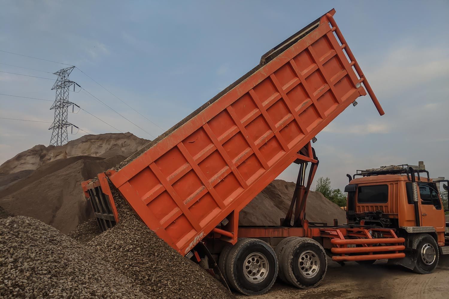 An orange dump truck discharging a load of gravel onto a large pile at an outdoor site with a utility pylon in the back.