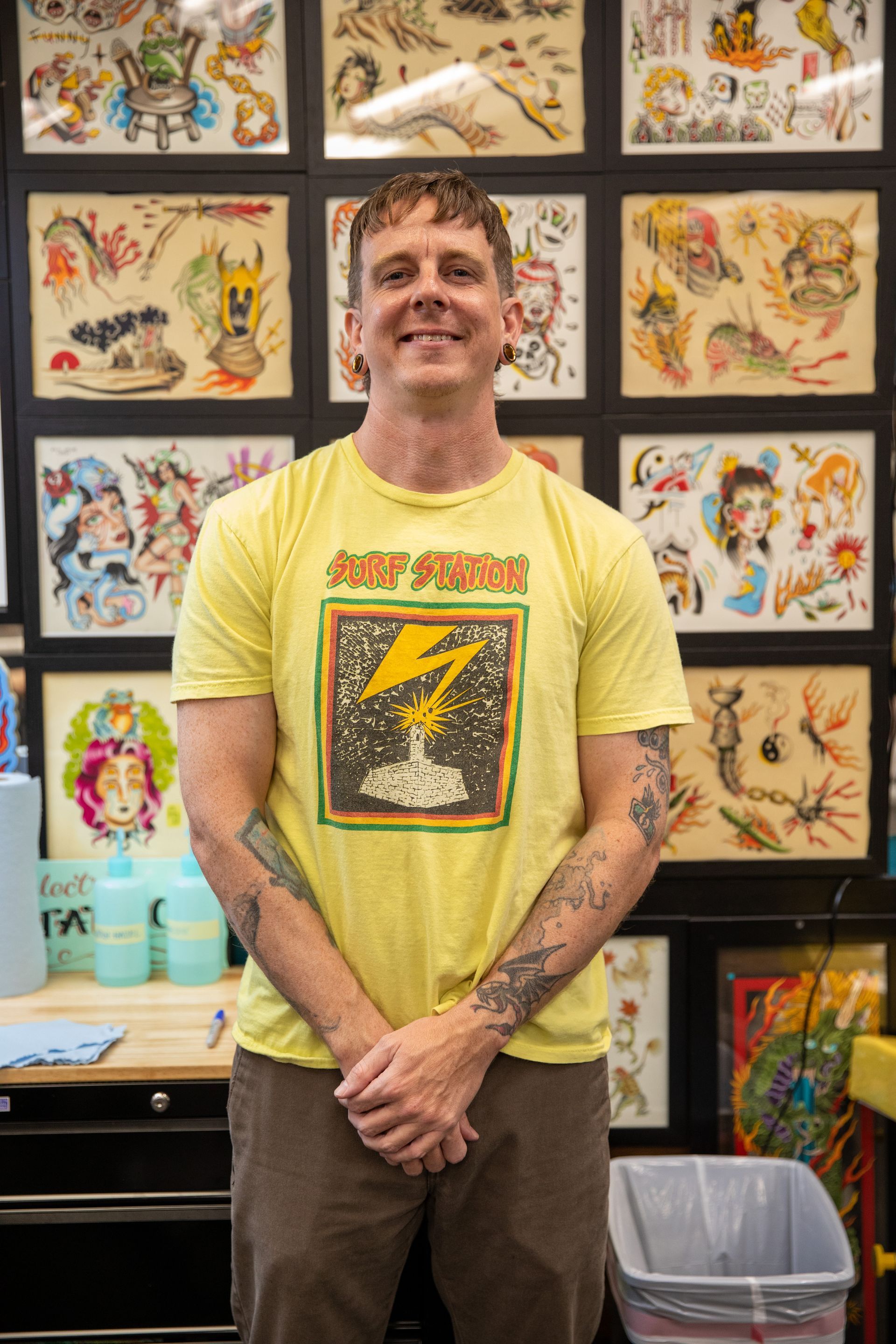 Person standing in a tattoo studio, smiling in a yellow graphic T-shirt with tattoo flash art on the wall behind.