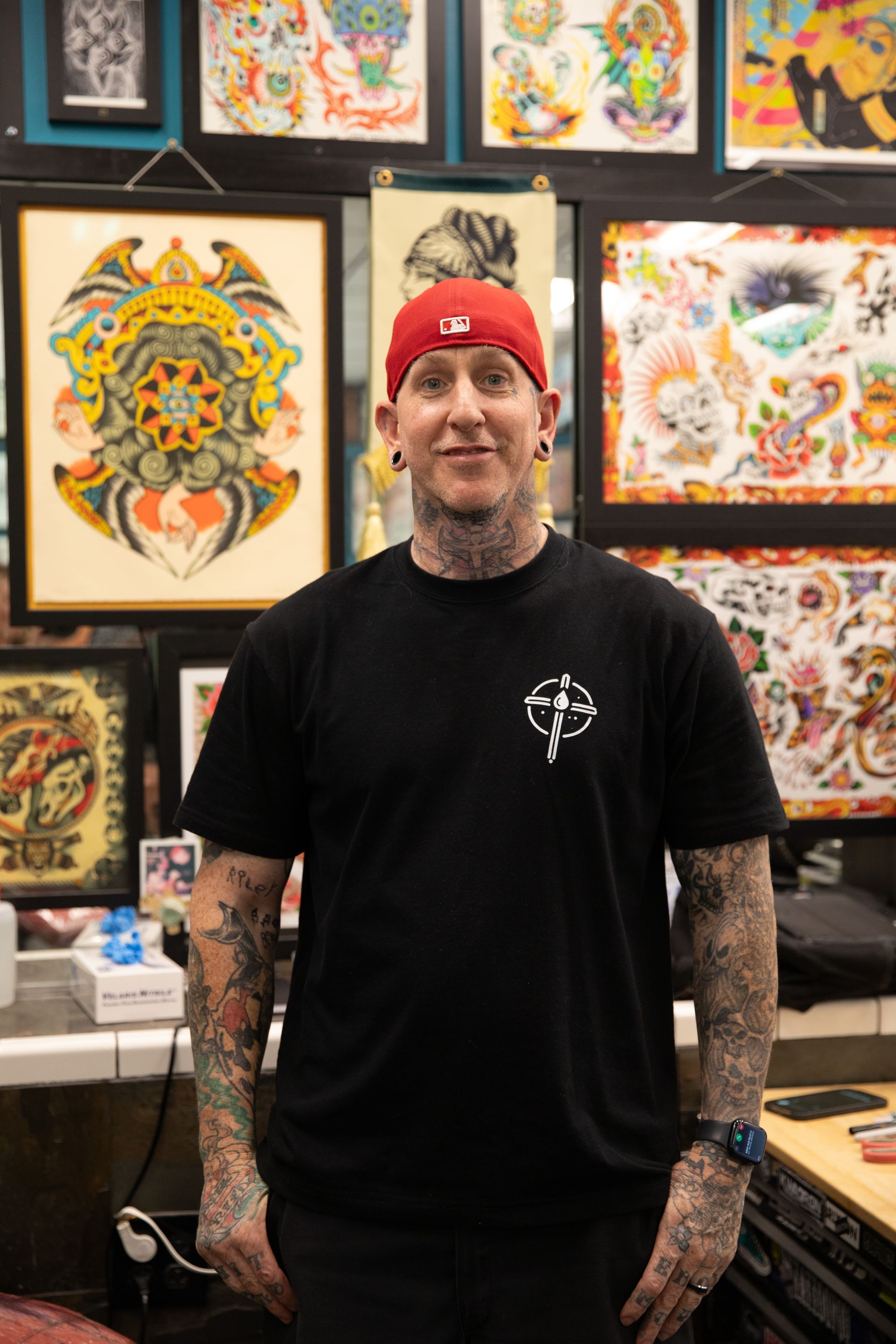 Man standing in a tattoo studio, wearing a black shirt and red beanie, with colorful tattoo art displayed behind him.
