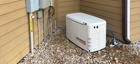 A standby generator installed outside a building. Beige siding, gray gravel, and electrical boxes are visible.
