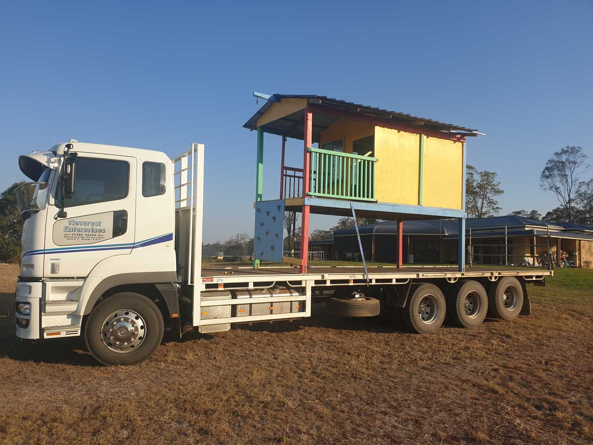 A white truck is carrying a small house on its flatbed.