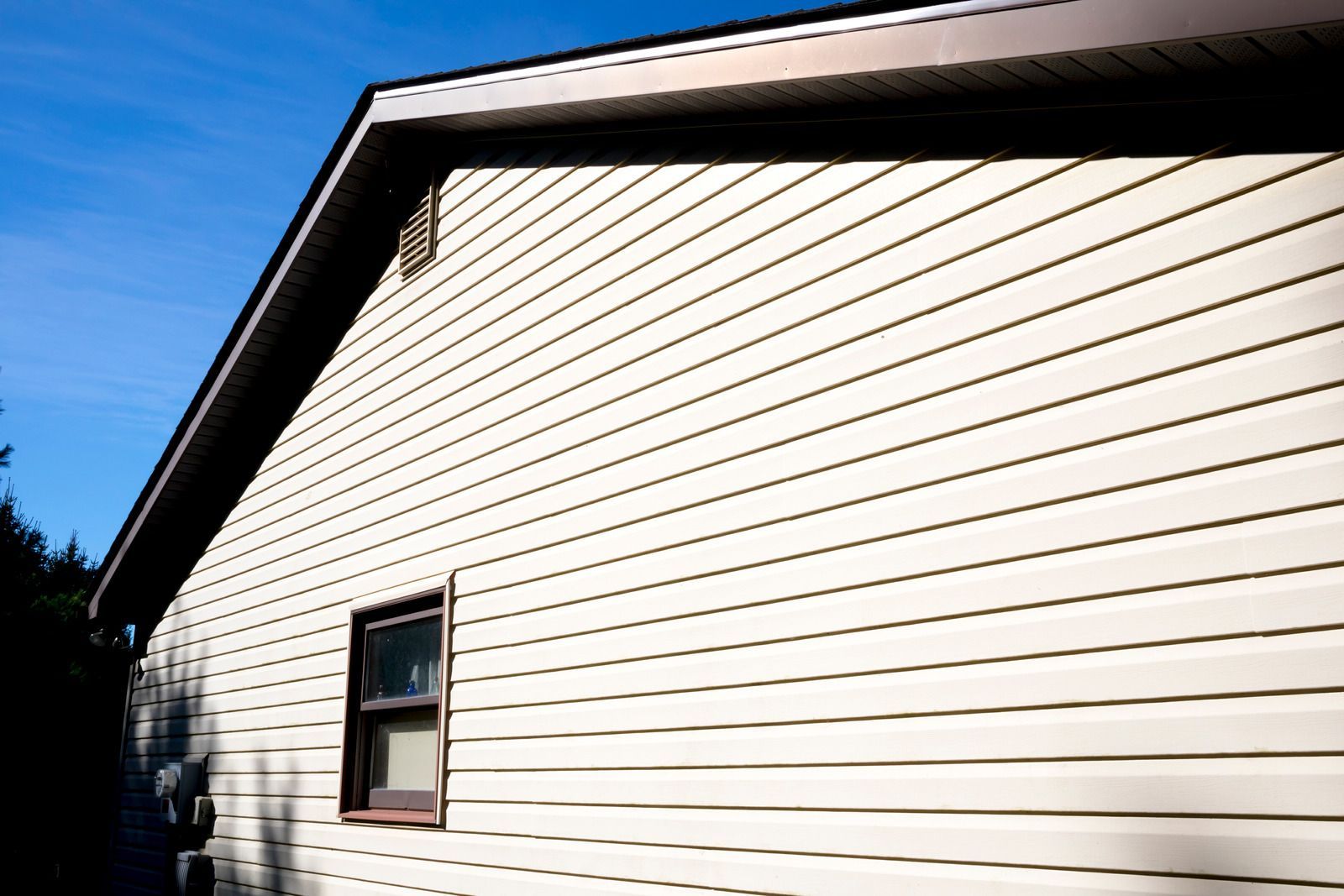 A side view of a house with beige horizontal siding, a single window, and a dark roof under a clear blue sky.