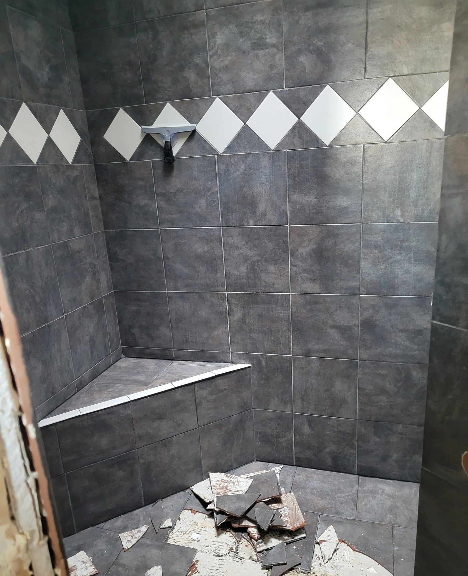 A shower stall with dark, textured square tiles, a white decorative diamond border, a corner seat, and construction debris.