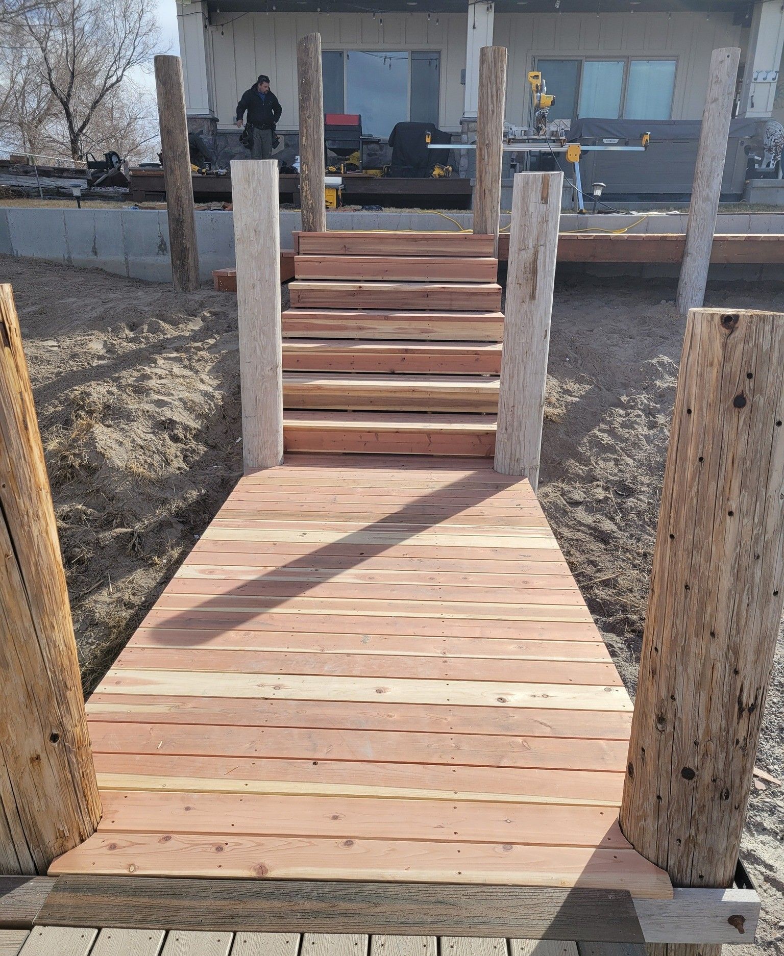 A wooden walkway with stairs leading up to a house under construction, framed by vertical rustic wooden posts.