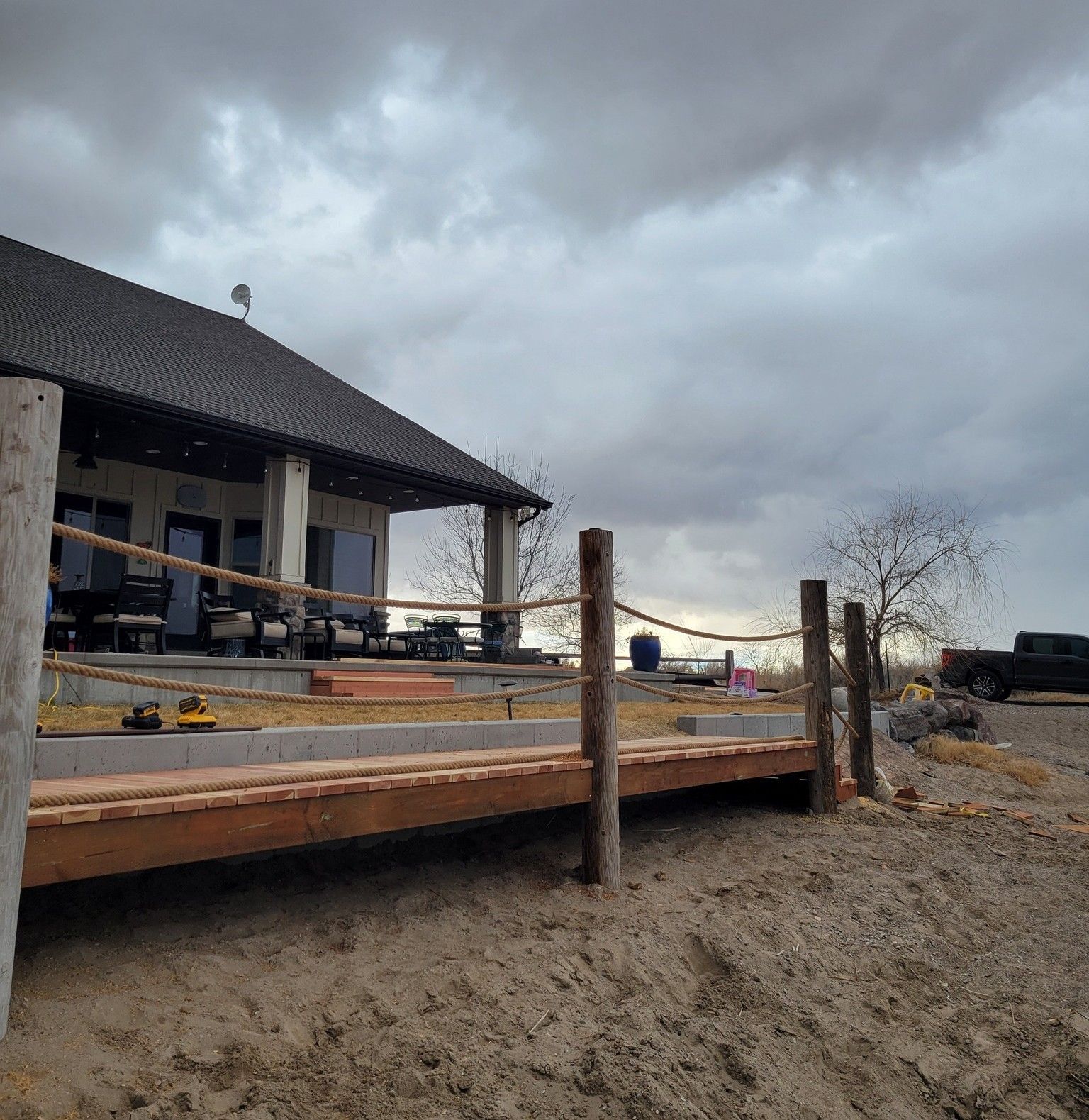 A wooden boardwalk with rope railings leads to a covered house patio under a cloudy, overcast sky.