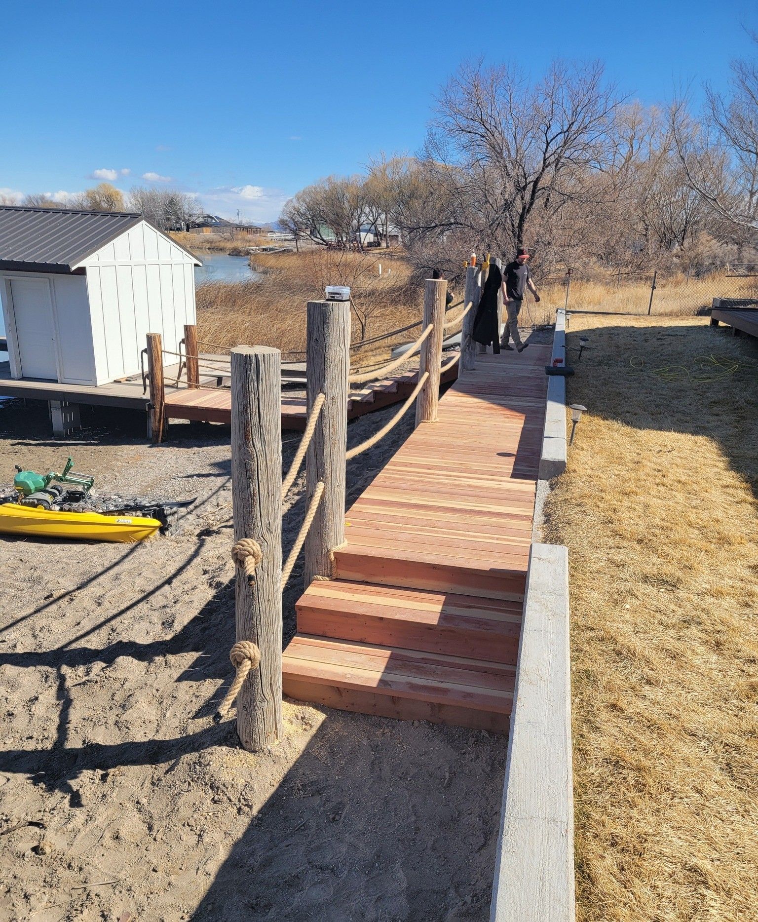 A wooden staircase and walkway with rope railings leads to a white shed by a sandy beach and water under a clear blue sky.