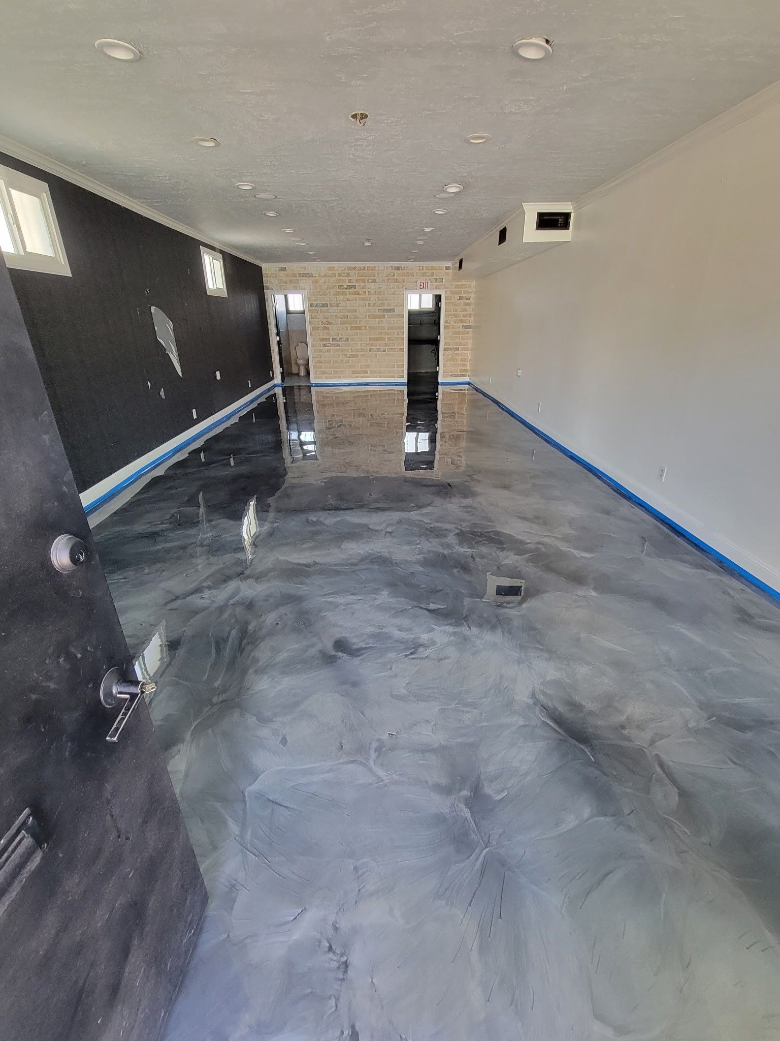 A long, narrow room with metallic grey epoxy flooring, a black wall on the left, a light wall on the right, and two doors.
