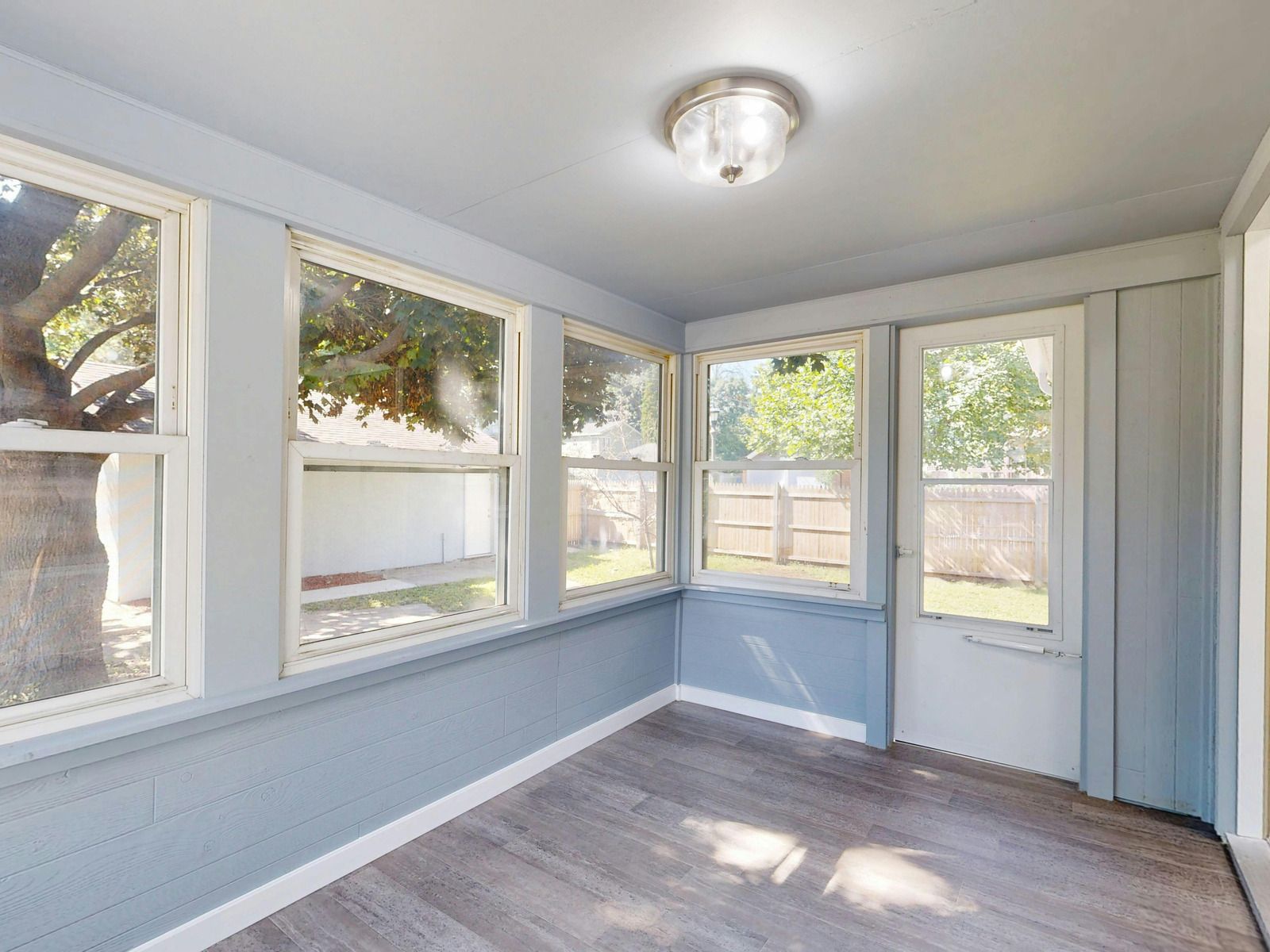 A sunroom with light blue walls, gray wood-look flooring, large windows, and a white door leading to a backyard.