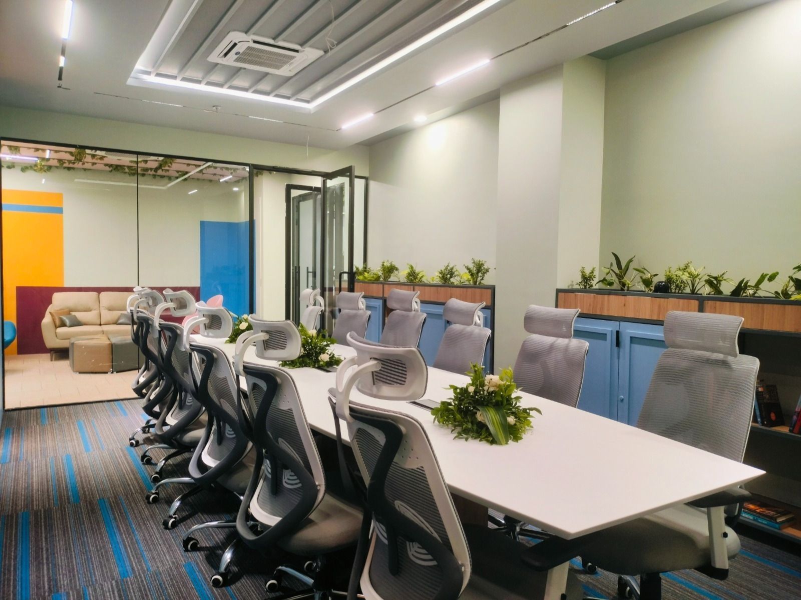 A long white conference table surrounded by grey mesh office chairs in a modern meeting room with light green walls.