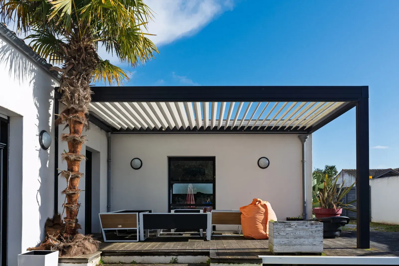 A modern white home with a black louvered pergola over a patio featuring outdoor seating and a palm tree under a blue sky.