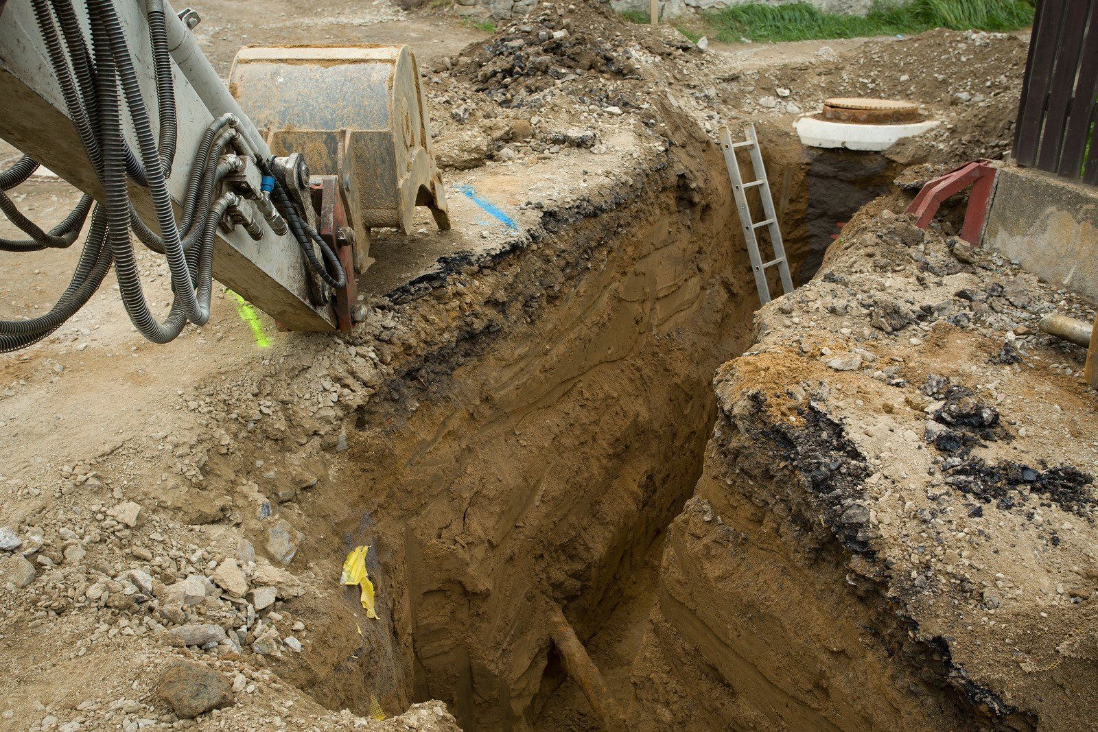 An excavator arm hangs over a deep, narrow construction trench with a metal ladder leaning against the side.