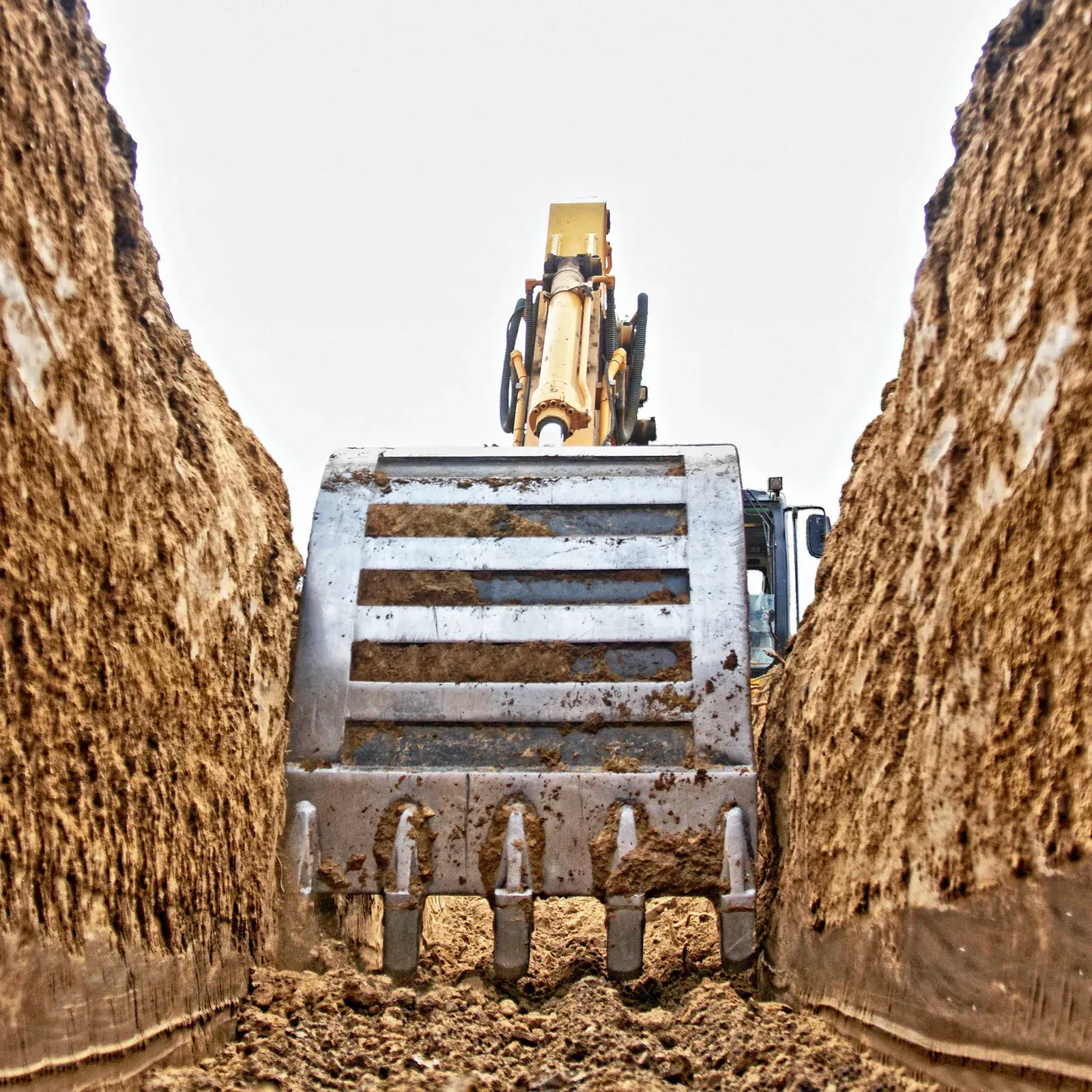A metal excavator bucket sits inside a narrow, deep trench dug into brown earth.