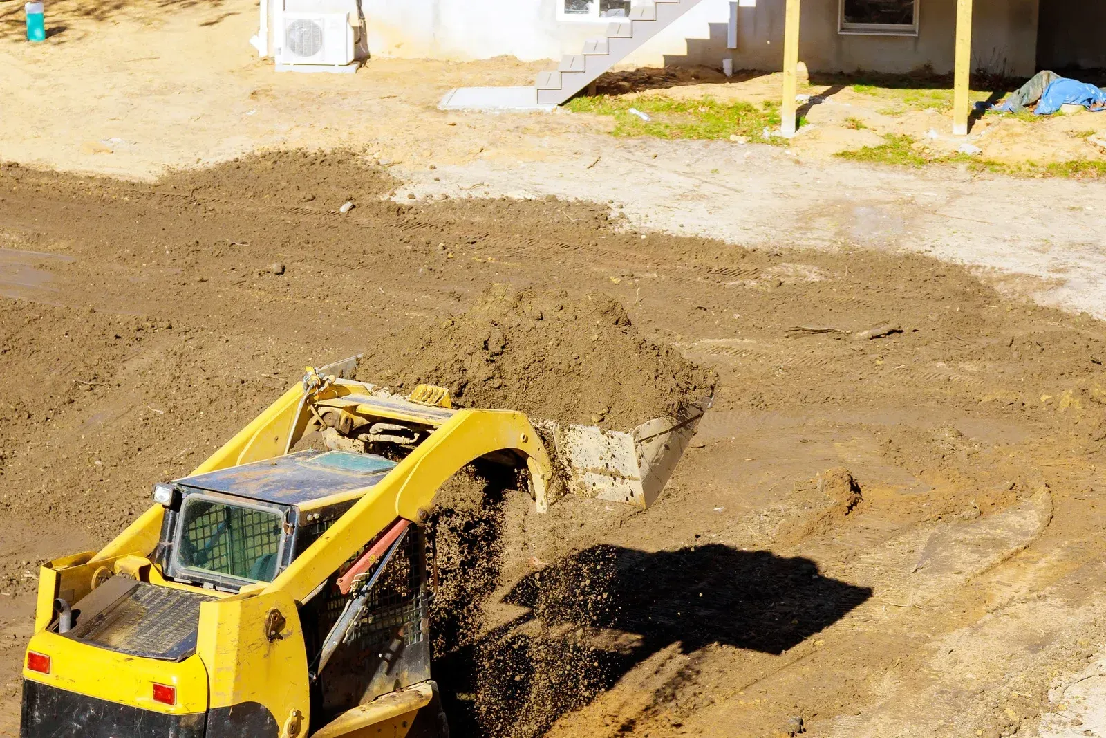 A yellow skid steer loader moves dirt on a construction site near a building with stairs.