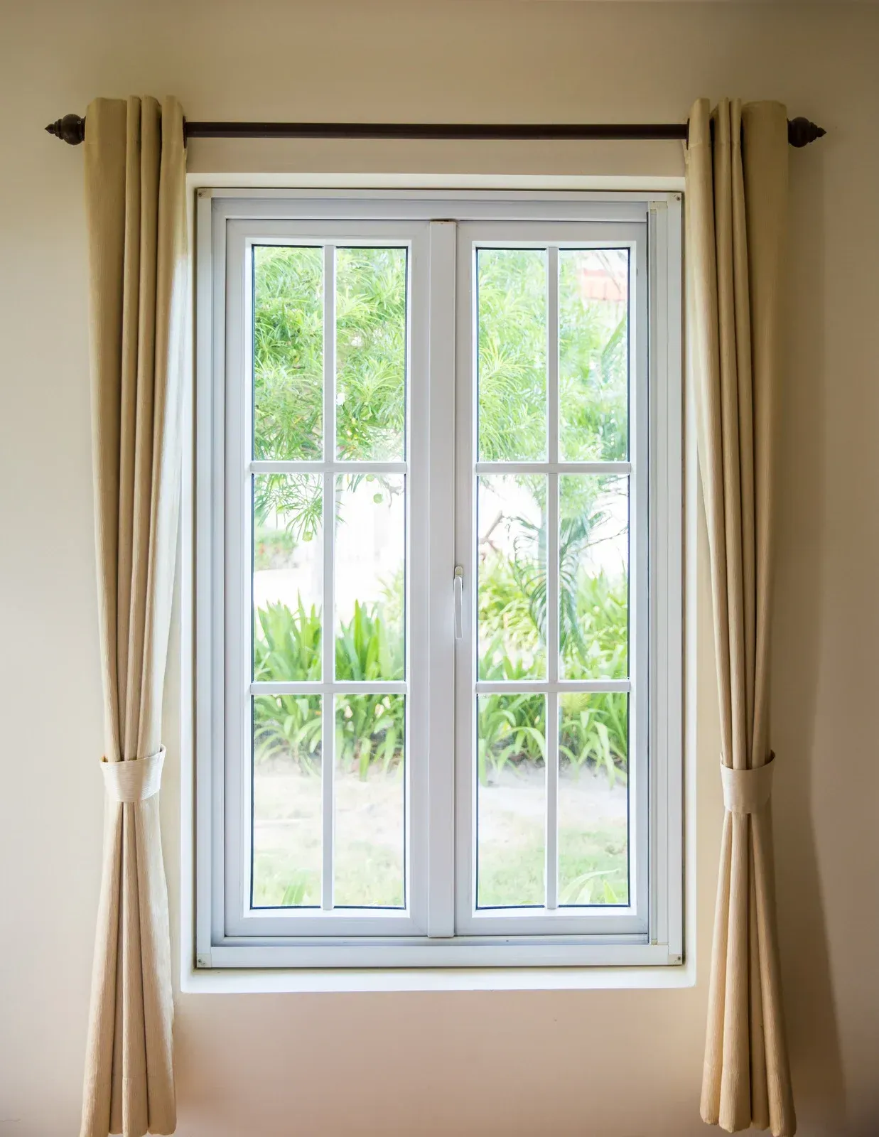 A white-framed window with six panes on each side, flanked by beige curtains on a dark rod against a light wall.