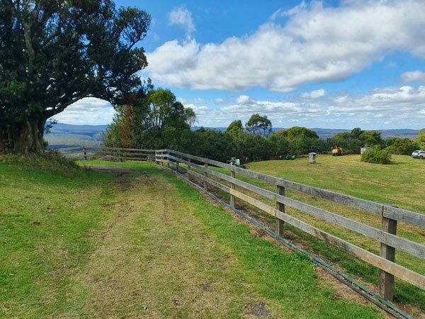 Grassy field with wooden fence, large tree, and distant hills under a cloudy blue sky.