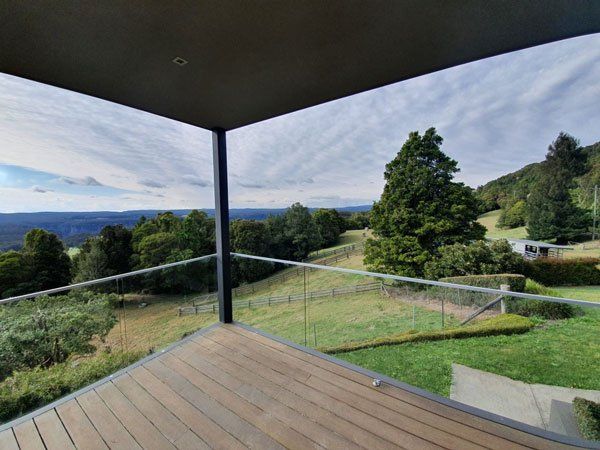 Balcony view: wooden deck, glass railing, overlooking green fields, trees, and cloudy sky.