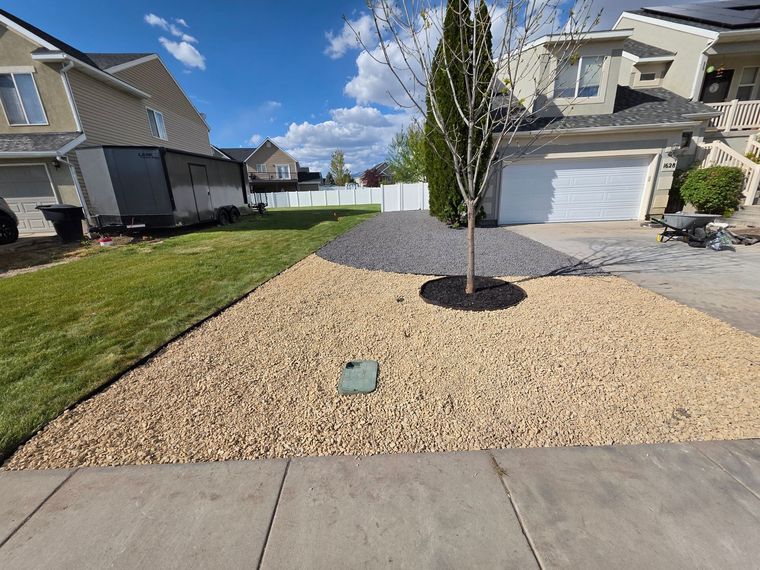 Front yard with gravel landscaping, a young tree, and a suburban house with a white garage door.