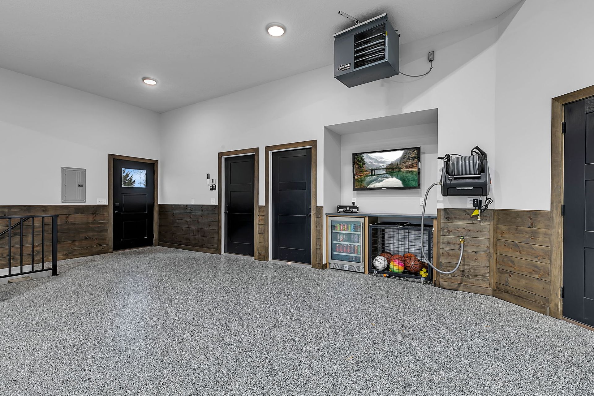 Garage interior with gray speckled floor, white walls, and dark doors and trim; a TV is mounted above a small fridge.