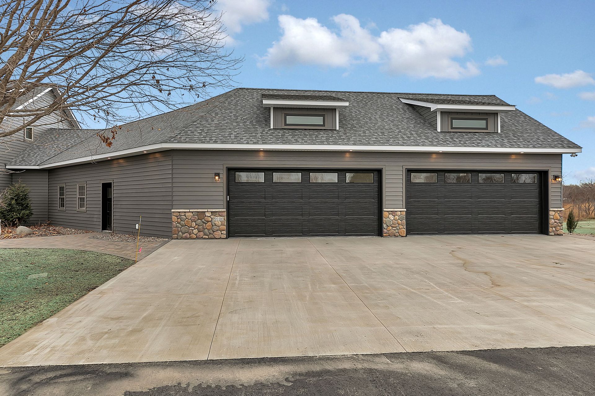 Gray garage with two bays, concrete driveway, and dormer windows under a cloudy sky.