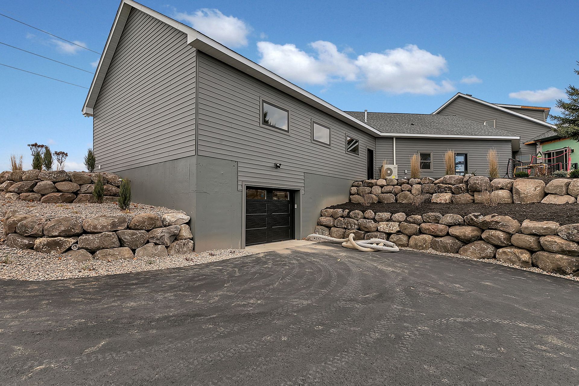 Gray building with a sloped roof and a black garage door. Stone walls border a paved driveway.