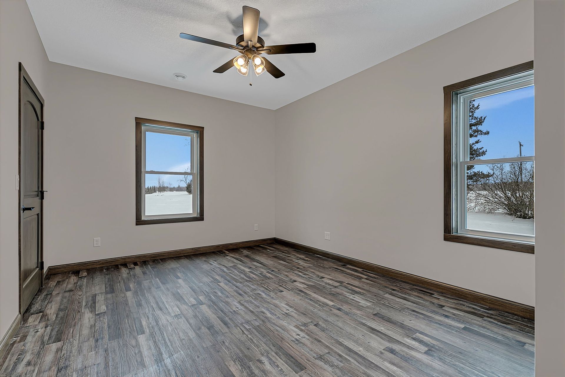 Empty room with gray walls, dark wood-look floor, windows with snowy view, ceiling fan.