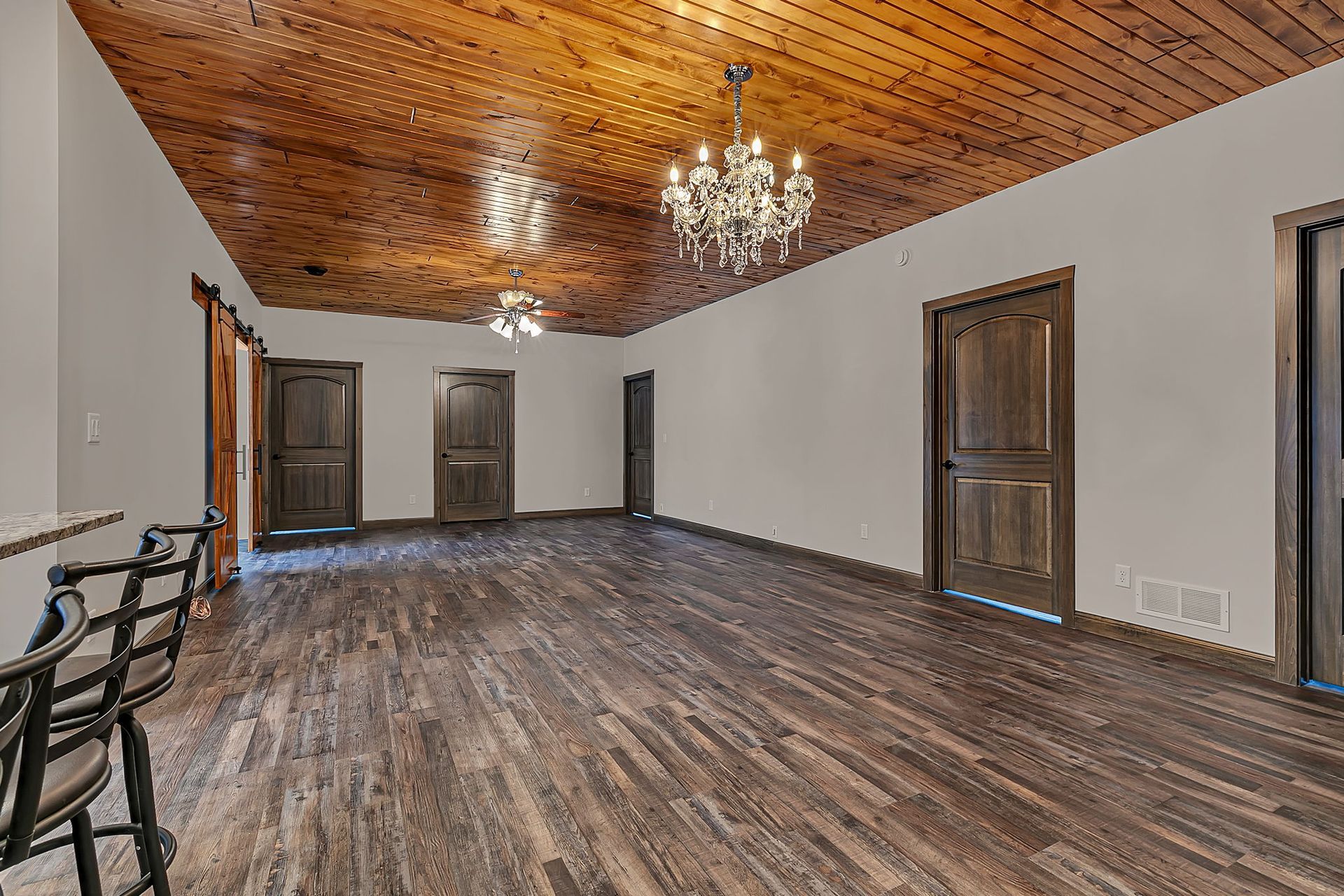 Empty room with wood floors and ceiling, white walls, dark doors, and chandeliers.