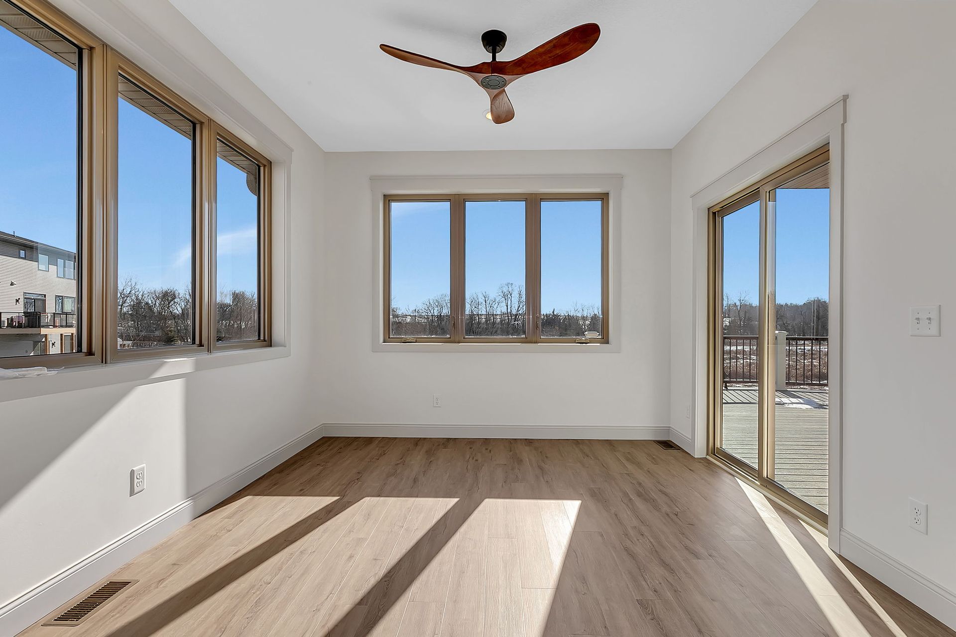 Empty room with large windows, sliding glass door, wooden floors, and a ceiling fan.