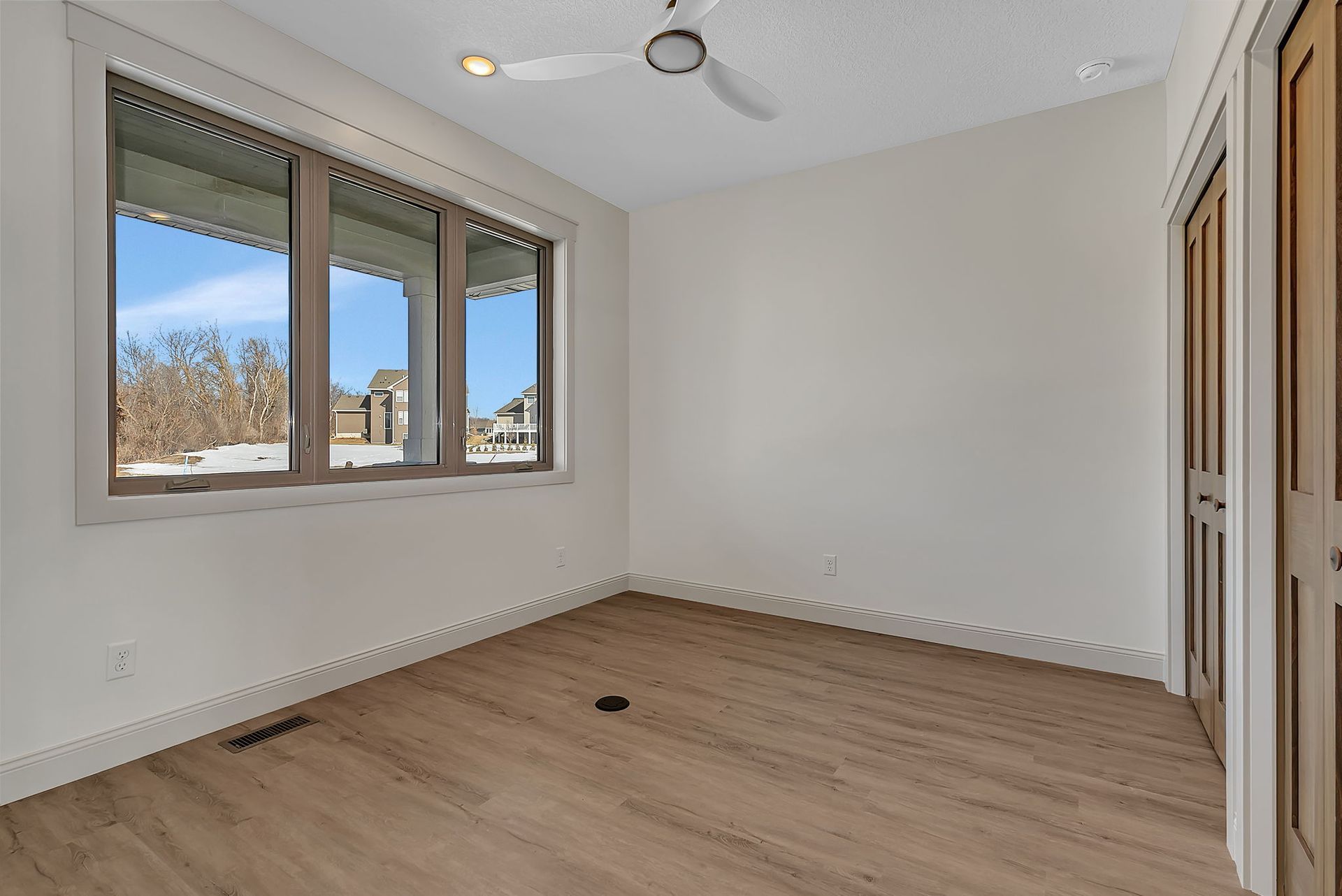 Empty room with light wood flooring, large window, and wooden door. Ceiling fan and white walls.