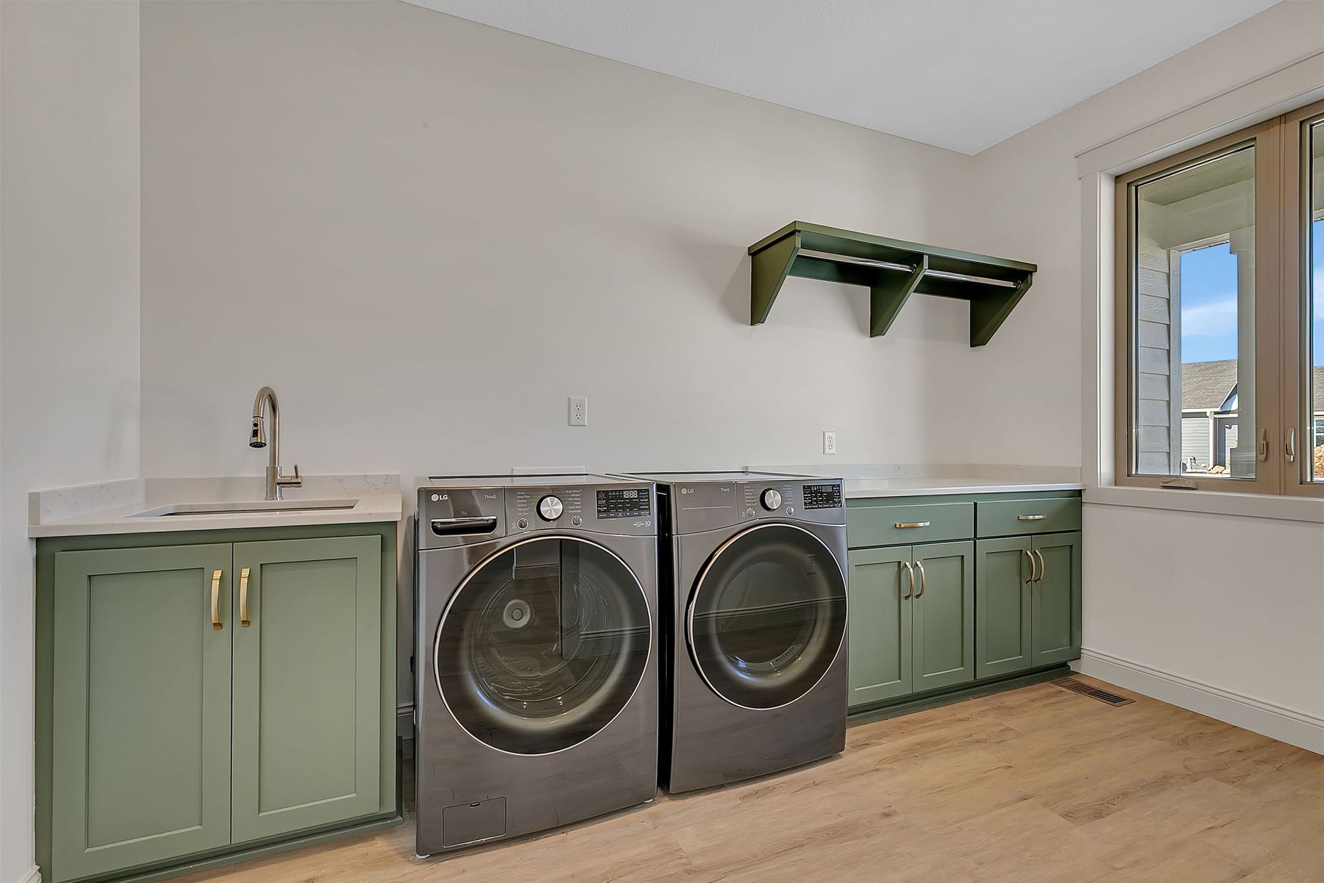 Laundry room with green cabinets, washer, dryer, sink, window, and wooden shelving.