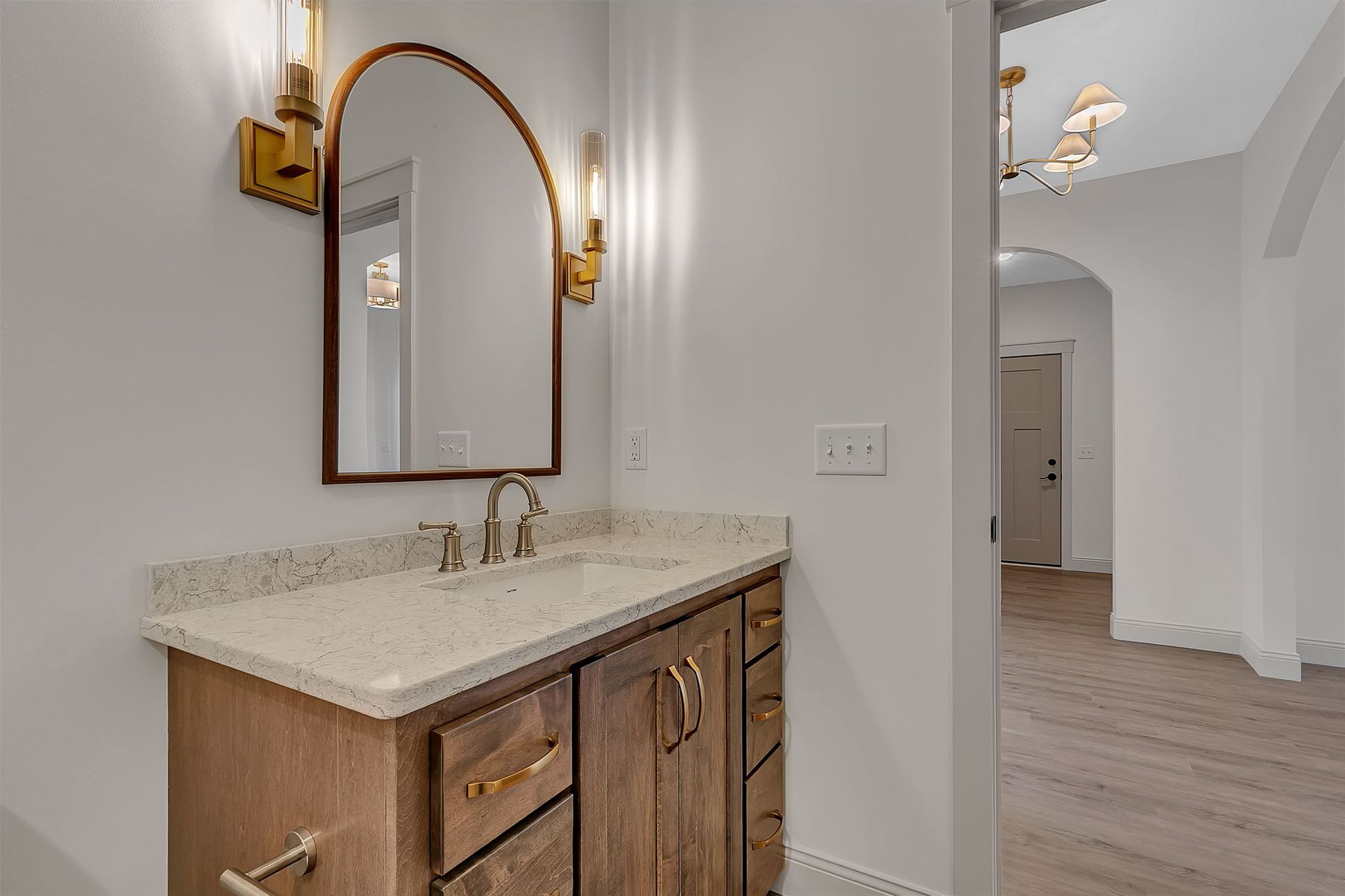 Bathroom with wooden vanity, arched mirror, gold sconces, and hallway view.