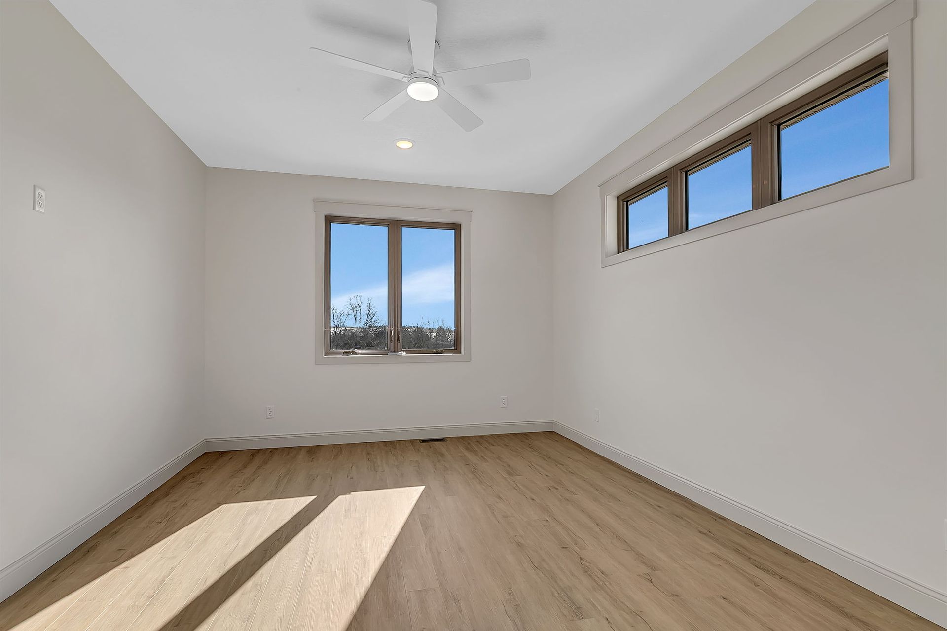 Empty room with light wood floors, white walls, and a window with a blue sky view. A ceiling fan is visible.